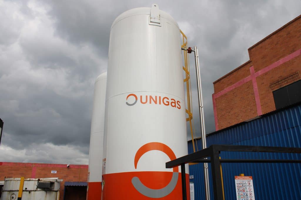 Tanque vertical blanco con el logo de Unigas junto a edificios de ladrillo, bajo un cielo nublado.
